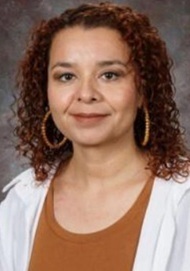A female teacher in a brown shirt and white jacket poses for a photo.