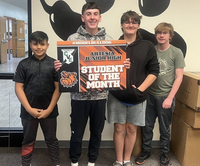 A teen boy in a grey sweatshirt posing with three other teen boys holds a sign reading "Student of the Month".