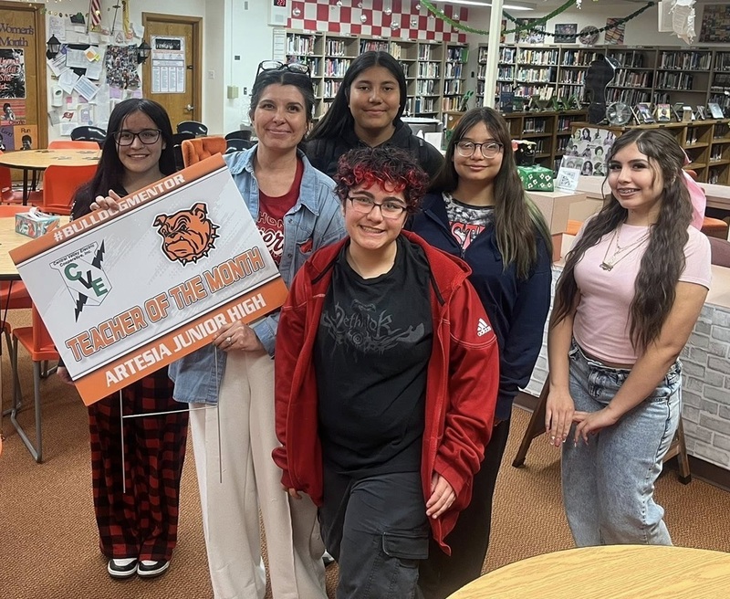 A female teacher in a jean jacket surrounded by five teen females holds a sign reading "Teacher of the Month" in a library.