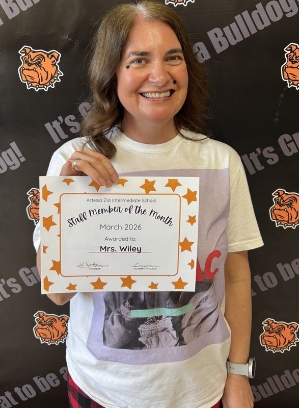 A female teacher in a white top holds a certificate reading "Teacher of the Month".