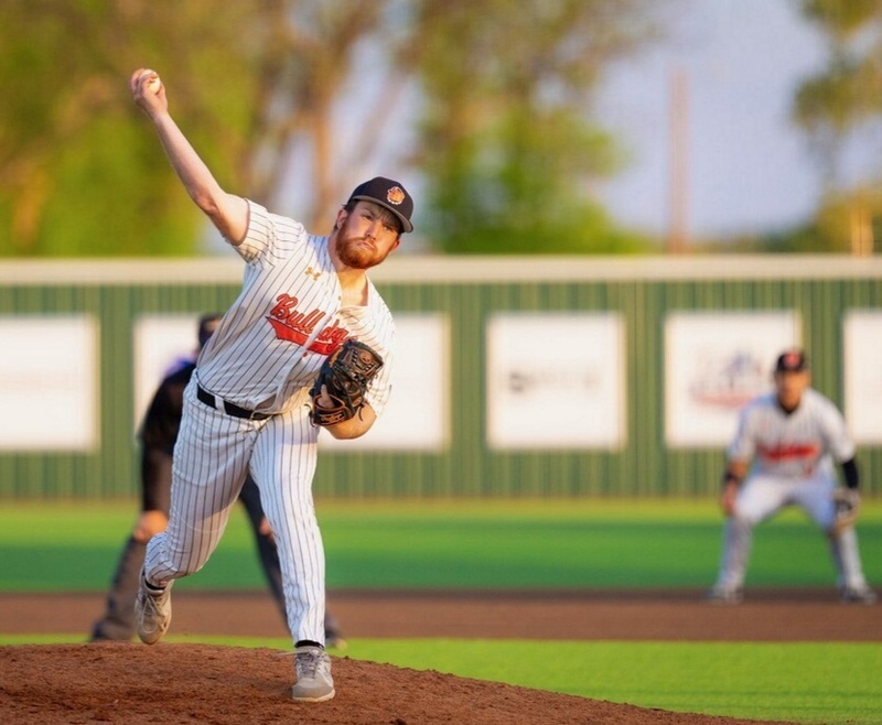 A teen boy in a white-and-black pinstriped baseball uniform with orange lettering prepares to release the ball from his right hand from the mound.
