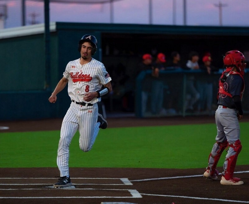 A teen boy in a white-and-black pinstriped uniform with orange lettering steps on home plate with his right foot as the opposing catcher in black, red and grey looks toward the field.