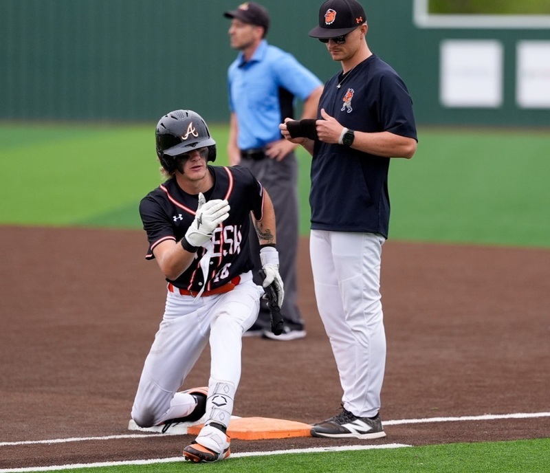 A teen boy in a black, white and orange baseball uniform gestures to the dugout from first base as a male coach watches.