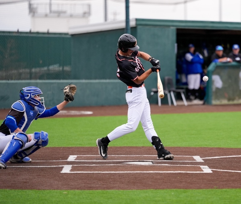 A teen boy in a black, white and orange baseball uniform in the left side of the batter's box swings as the ball comes into view. A catcher in blue and black is also visible.