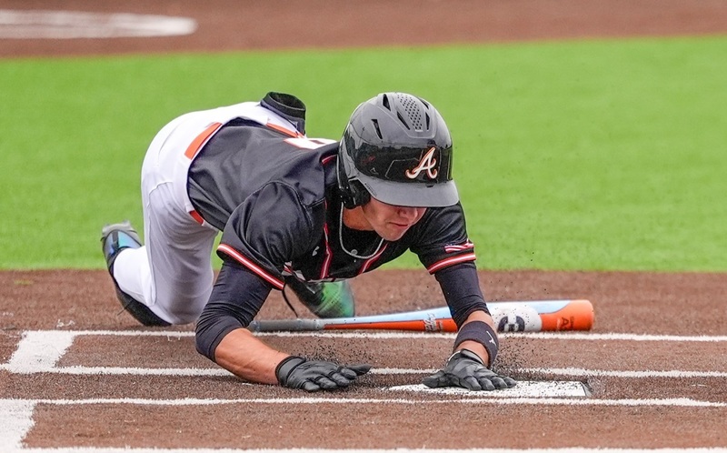 A teen boy in a black, white and orange baseball uniform and black batting helmet slides over top of an orange-and-white bat and onto home plate.