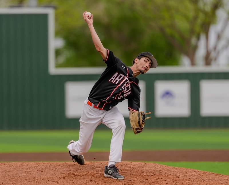 A teen boy in a black, white and orange baseball uniform prepares to sling the ball from his right hand from atop the mound.
