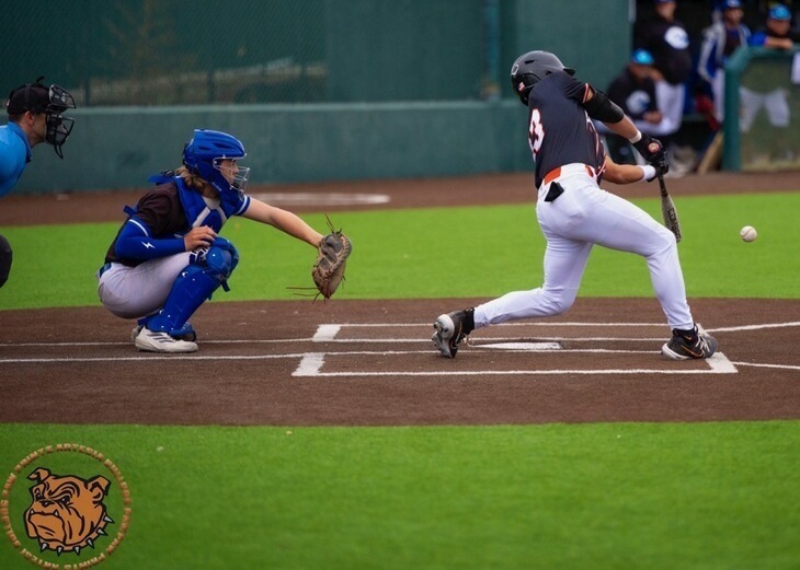 A teen boy in a black, white and orange baseball uniform swings from the right side of the batter's box as the ball comes in knee-high. A catcher in blue and black is also visible.