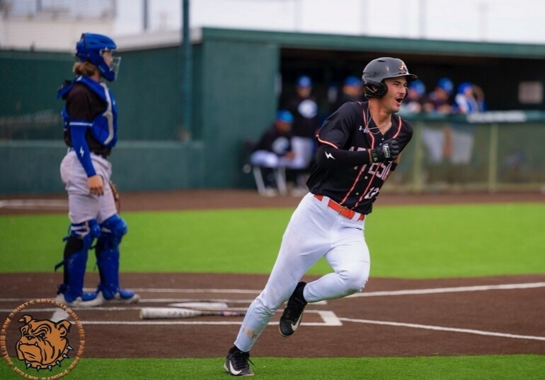 A teen boy in a black, orange and white baseball uniform watches his single with a smile on his face as he runs toward first base. A catcher in black and blue also stands behind home plate and watches.