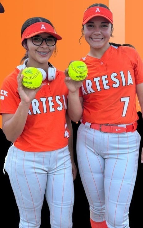Two teen girls in orange, white and grey softball uniforms smile for the camera while holding softballs.