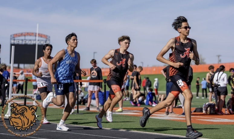 Four teen boys, two in black and orange track uniforms, one in blue, and one in white, run along a track.