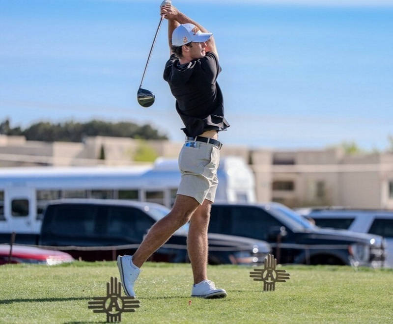 A teen boy in a back shirt, white cap and khaki shorts tees off on a golf course.