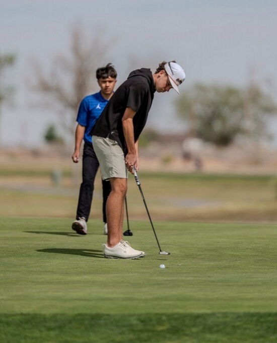 A teen boy in a black shirt, white cap and khaki shorts putts the ball. Another golfer in a blue shirt and black pants looks on.