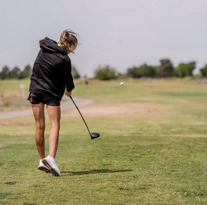 Seen from behind, a teen girl in a black hoodie and black skirt tees off. The ball can be seen flying up the fairway in front of her.