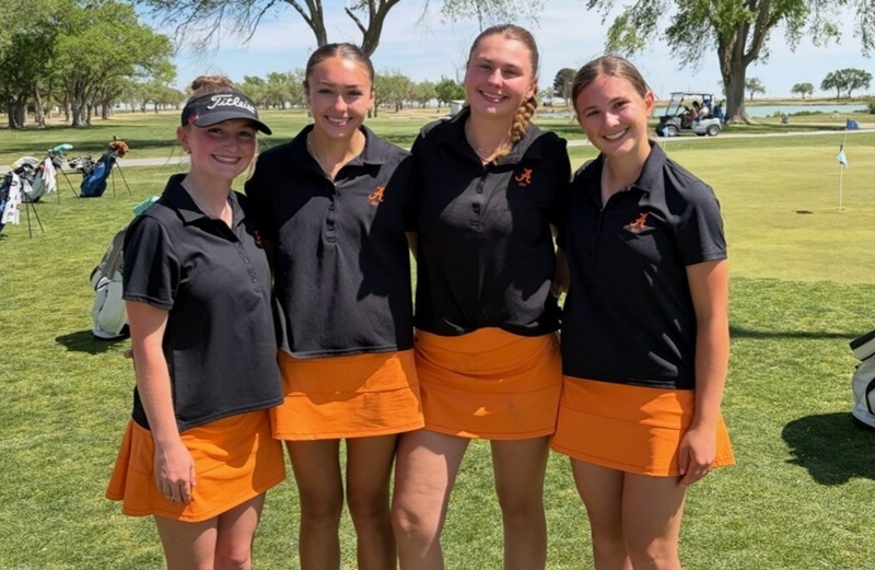 Four teen girls in black polo tops and orange skirts pose for a photo on a golf course.