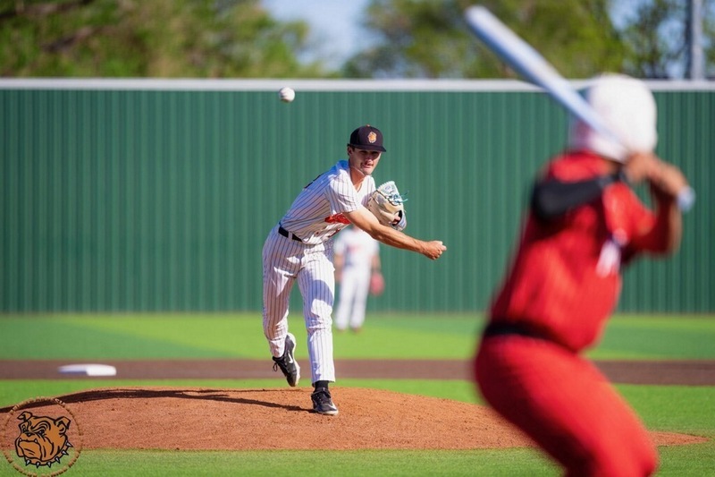 A teen boy in a pin-striped baseball uniform with orange lettering throws a pitch toward a batter in all red from the mound.