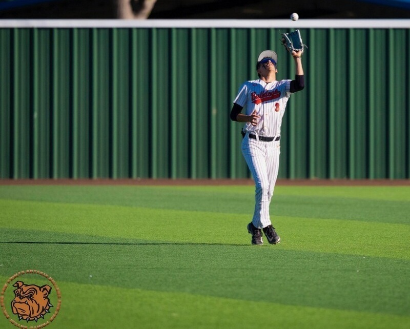 A teen boy in a pinstriped baseball uniform with orange lettering watches the baseball into his glove, held above his head with his left hand.