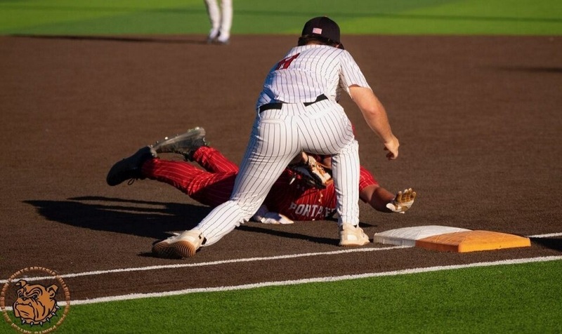 A teen boy in a pinstriped baseball uniform with orange lettering tags a runner in all red out as he attempts to dive back into first base.