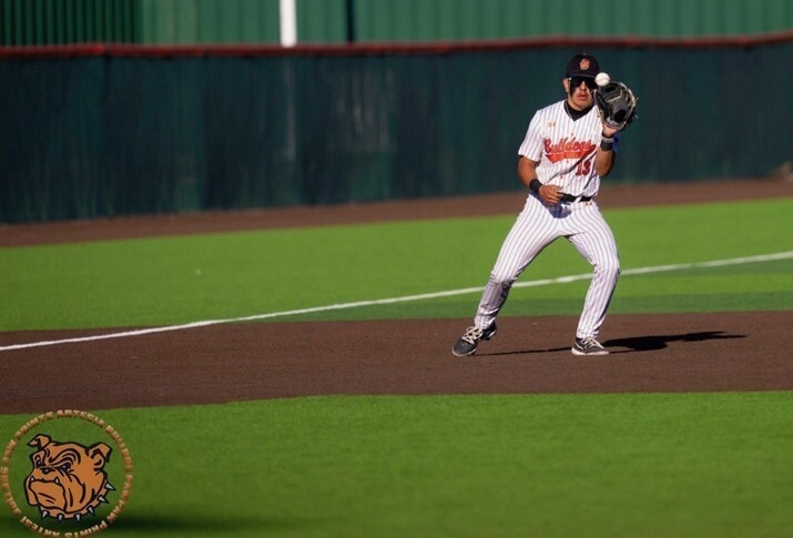 A teen boy in a pinstriped baseball uniform with orange lettering watches the baseball into his glove, held at eye level with his left hand.