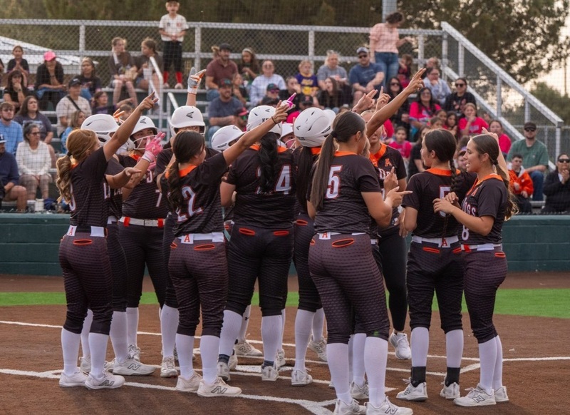 A girls' softball team in black and orange uniforms celebrate at home plate.