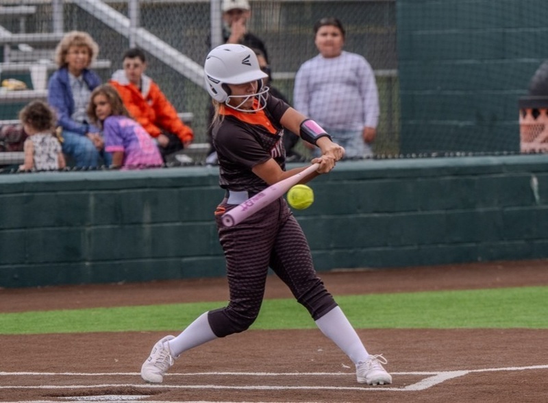 A teen girl in a black and orange softball uniform connects the bat with the ball.