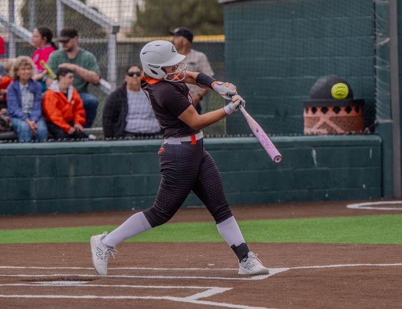 A teen girl in a black and orange softball uniform swings as the ball heads toward her bat.