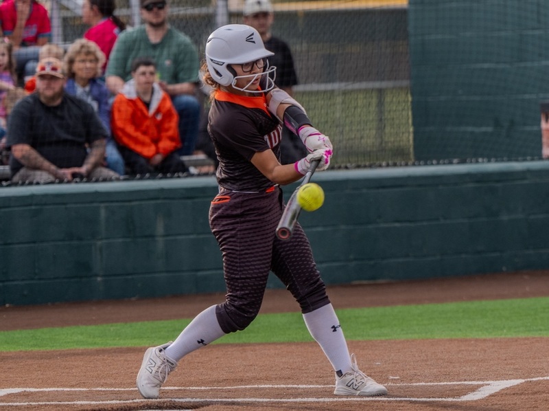 A teen girl in a black and orange softball uniform connects with the ball.
