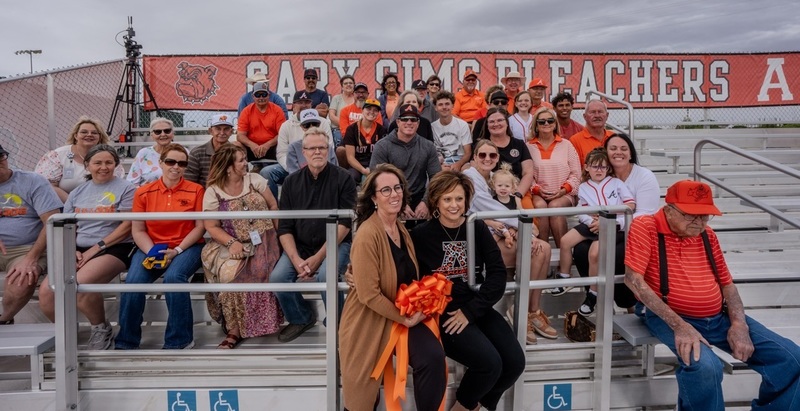 A group of men, women and children pose for a photo while seated in a set of bleachers. A sign behind them reads "Gary Sims Bleachers".