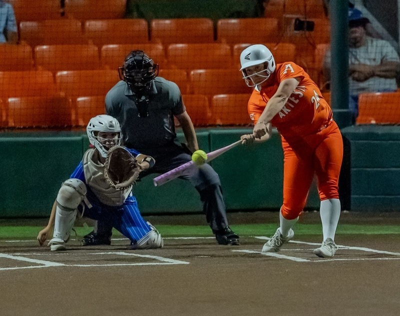 A teen girl in an orange softball uniform connects with the ball. A catcher in blue and an umpire are also visible. 