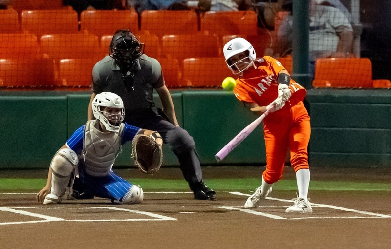 A teen girl in an orange softball uniform swings as the ball heads toward her bat. A catcher in blue and an umpire are also visible.