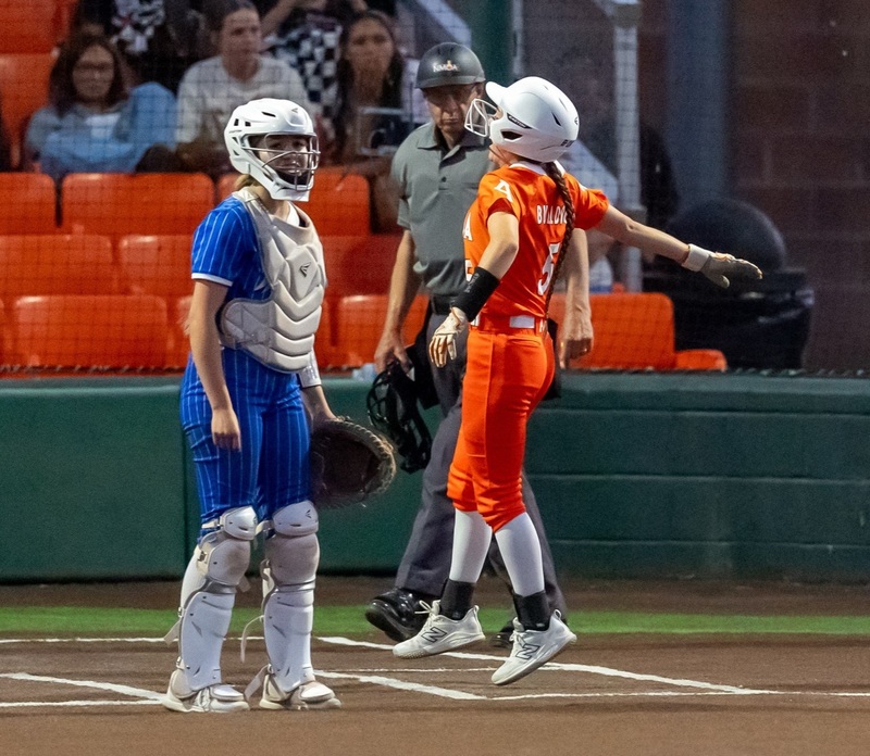 A teen girl in an orange softball uniform prepares to step on home plate as a catcher in blue looks out toward the field.
