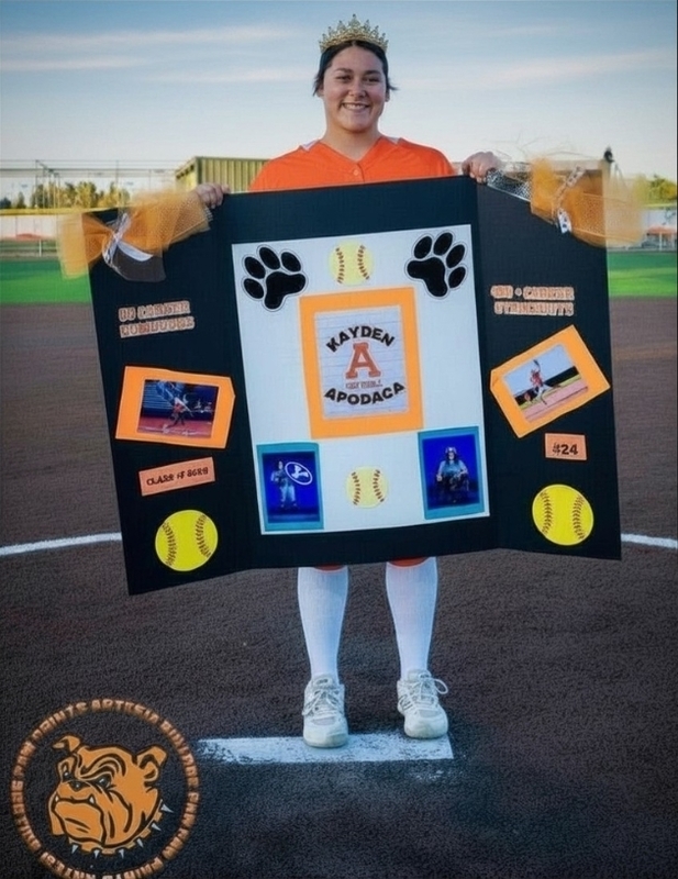 A teen girl in an orange softball uniform smiles as she stands on the pitching rubber holding a sign with softball and paw graphics.