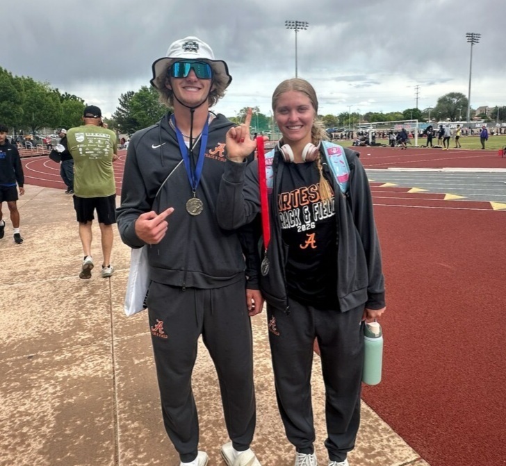 A teen boy and teen girl in black track warm-ups smile for the camera while standing alongside a red track surface.