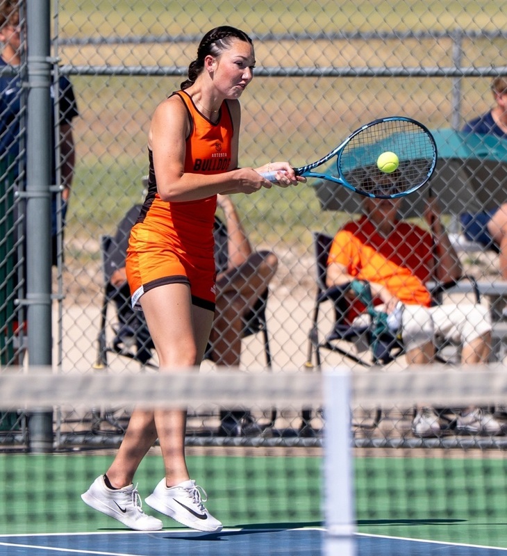 A teen girl in an orange tennis uniform hits the ball back toward the net with her racket. 