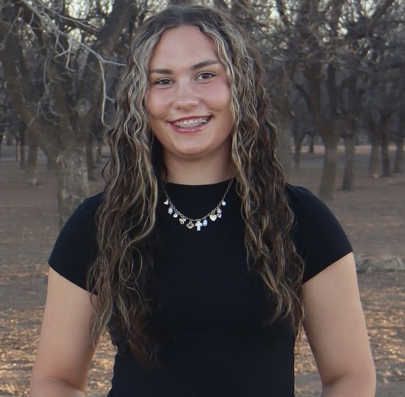 A teen girl with long dark hair tinted blonde and a black top smiles for the camera while standing in front of a grove of trees.