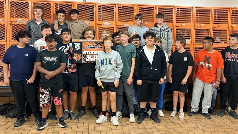 A teen boy surrounded by other teen boys stands in front of a wall of orange lockers and holds a sign reading "Student of the Month".