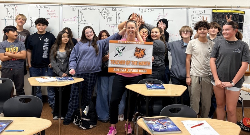 A female teacher surrounded by teen students, several of whom are giving her bunny ears, holds a sign reading "Teacher of the Month".