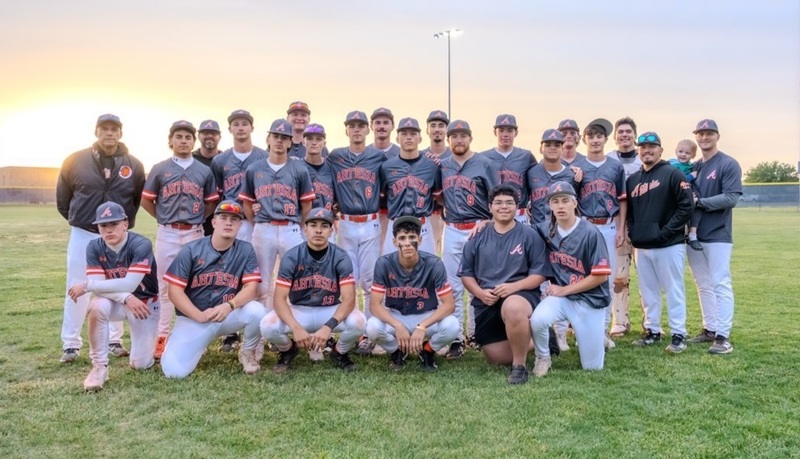 A high school baseball team in grey, orange and white uniforms poses for a photo in the outfield grass.