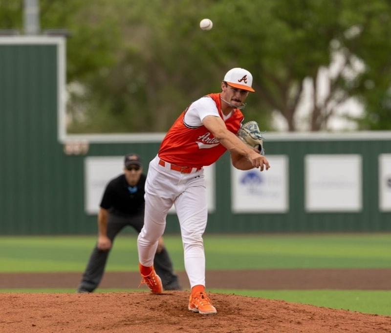A teen boy in an orange and white baseball uniform throws a pitch from atop the mound.