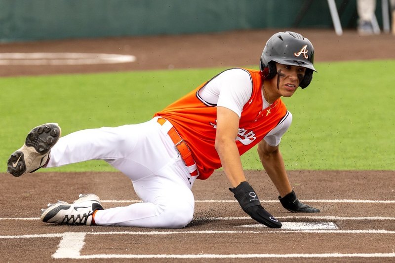 A teen boy in an orange and white baseball uniform slides across home plate.