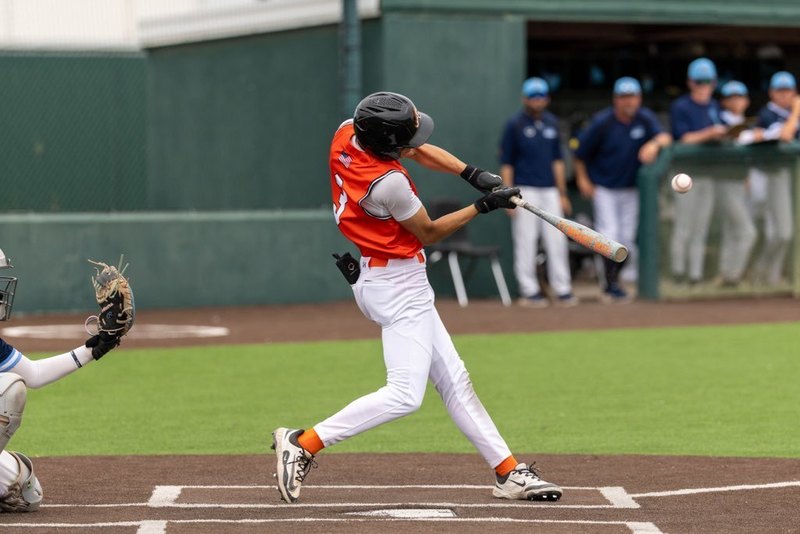 A teen boy in an orange and white baseball uniform connects with the ball at the plate.