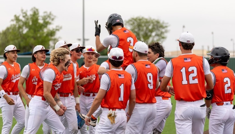 A teen boy in an orange and white baseball uniform is lifted up into the air by a large group of teammates.