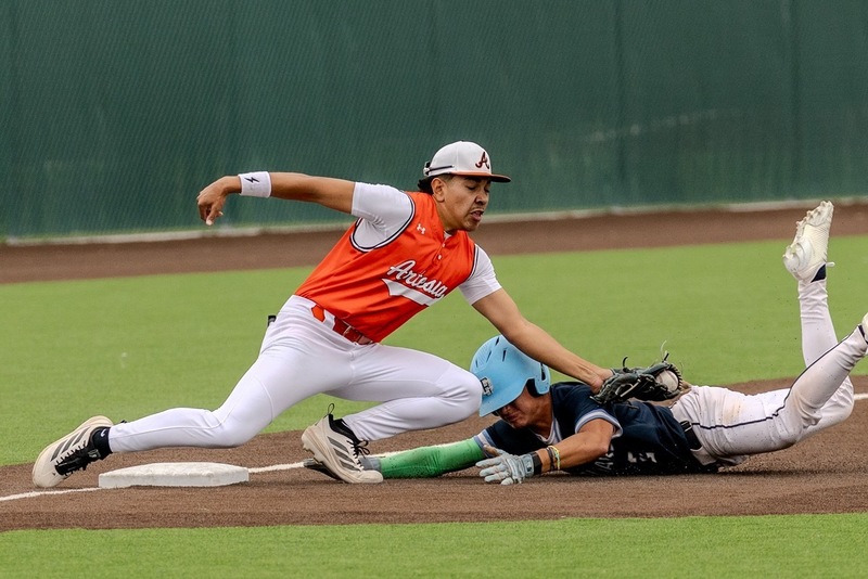 A teen boy in an orange and white baseball uniform tags an opponent in baby blue out as he attempts to slide into third base.