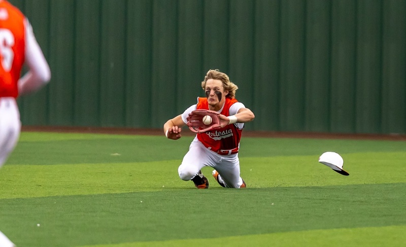 A teen boy in an orange and white baseball uniform dives forward to catch the ball. His hat is seen flying off.