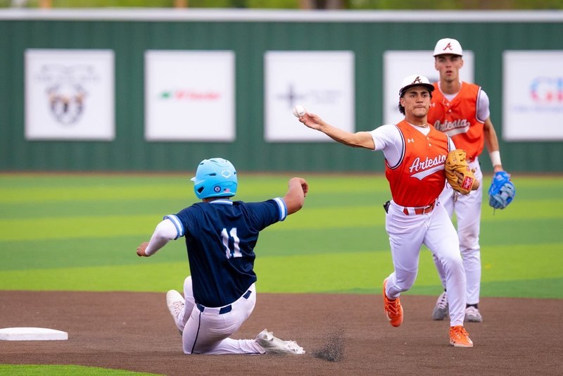 A teen boy in an orange and white baseball uniform throws to first base as an opponent in navy and baby blue is seen sliding toward second.