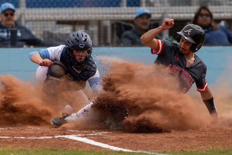 A teen boy in a black and orange baseball uniform sends large clouds of dust flying as he slides into home plate in front of a catcher in white and baby blue.