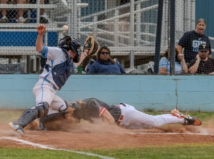 A teen boy in a black and orange baseball uniform is seen sliding head first into home plate behind a catcher in white and baby blue. 