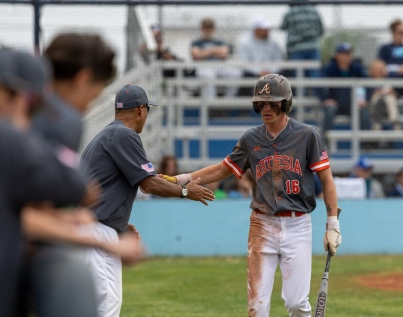 A teen boy in a black and orange baseball uniform is congratulated by a male coach as he walks back to the dugout.