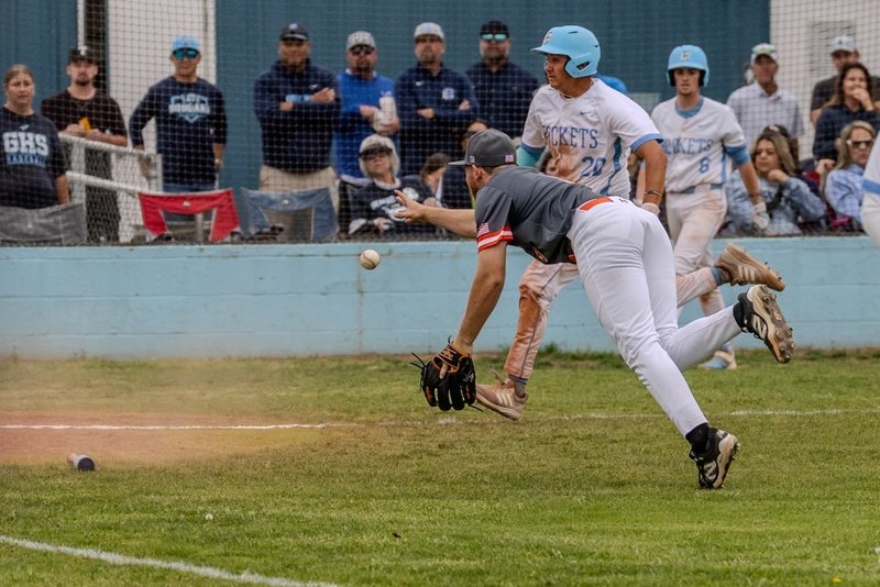 A teen boy in a dark grey and orange baseball uniform lunges toward home plate as he flips the ball out of his right hand. A baserunner in white and baby blue is seen attempting to beat the throw.