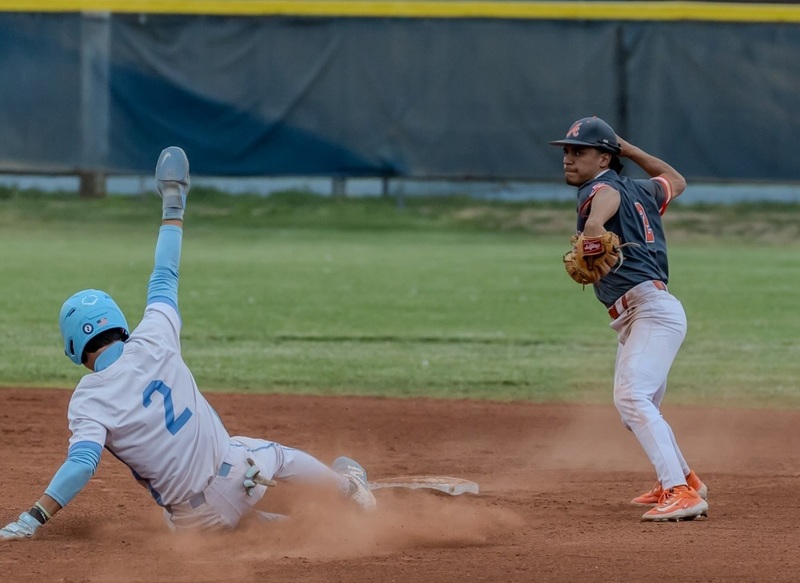 A teen boy in a dark grey and orange baseball uniform prepares to throw the ball to first as an opponent in white and baby blue slides toward second base.