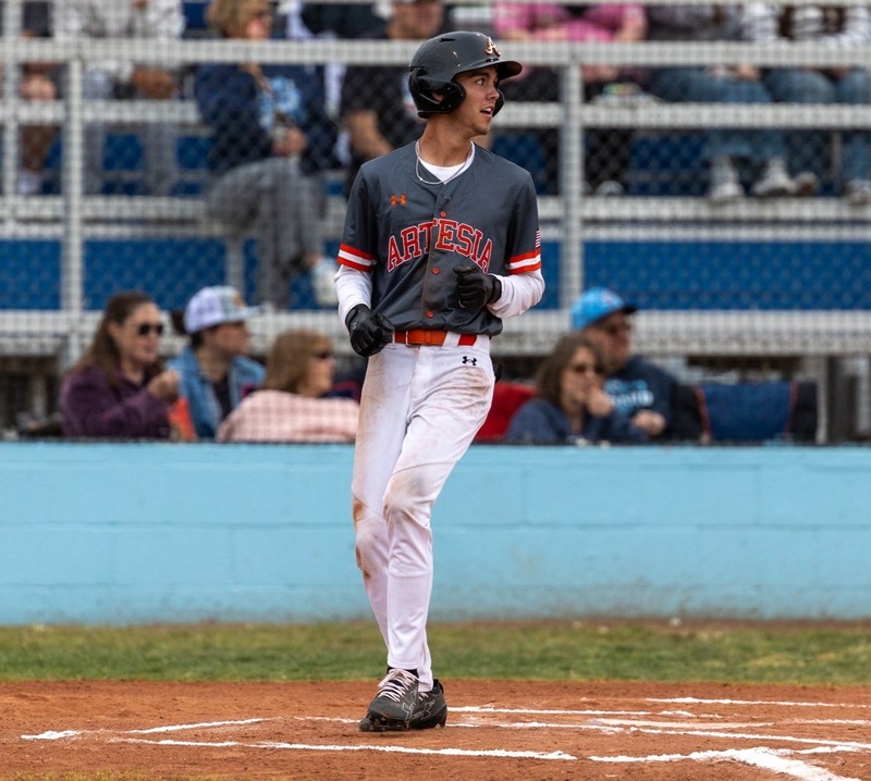 A teen boy in a dark grey and orange baseball uniform looks toward the field as he steps on home plate.